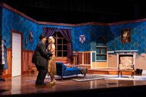 A theatrical stage set with actors in a vintage-style room featuring a chaise lounge, blue walls, and wood paneling.