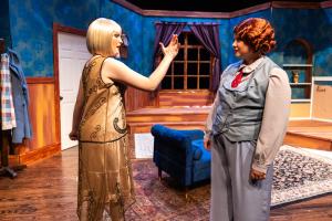 Two actors on a stage set resembling a living room, with one gesturing towards the other.