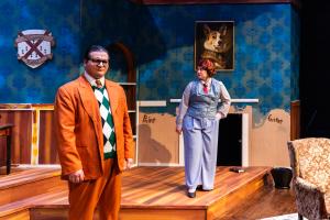 Two actors on a stage with a patterned blue wall and various props, including a coat of arms and a painting.