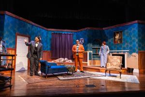 Actors on stage in a dramatic room setting with blue walls and elegant furnishings.