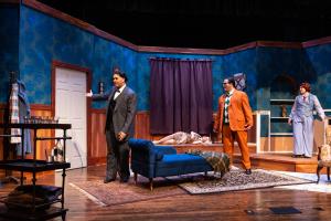 Theatrical stage set with blue walls, a blue chaise lounge, and actors in colorful costumes.