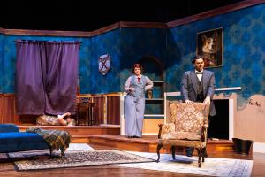 Theatrical stage set resembling a vintage living room with actors, featuring blue walls, a purple curtain, and period furniture.
