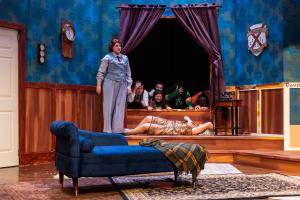 A stage play set with a blue chaise longue, a woman standing, and a person lying on the floor. Several people peer from behind a curtain.