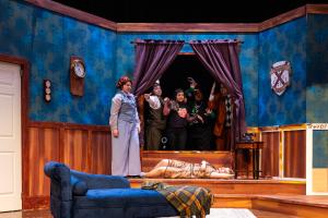 Theatrical stage set depicting a group of people in a living room, with one person lying on the floor.