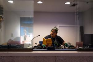 Person reading a book behind a glass partition with electronic devices and a microphone in front.