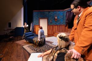 Actors perform on a stage set resembling a living room with blue wallpaper and wooden paneling.