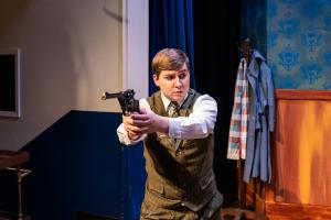 An actor on stage holding a prop gun, dressed in vintage attire with a backdrop of a blue curtain and wooden paneling.