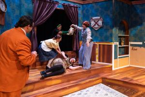 A dramatic stage scene with five actors in a richly decorated room, featuring blue walls and wooden flooring.