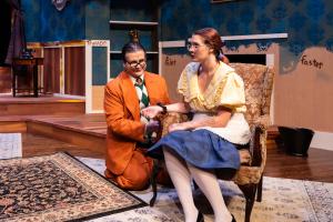 A man in an orange suit kneels next to a woman seated in an armchair during a theatrical performance.