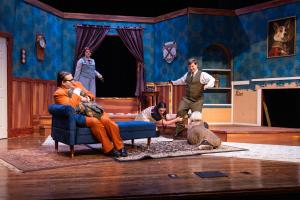 Theater stage set of a vintage parlor with actors in various poses and costumes.