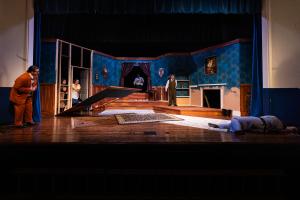 Theatrical stage set depicting a room with blue wallpaper, a raised floor section, and two actors in suits.