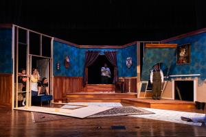 Theatrical stage set depicting a room with blue wallpaper, a raised floor section, and two actors in suits.