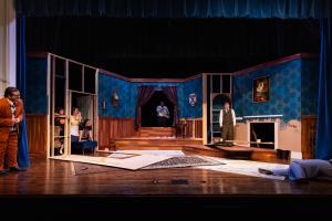 Theatrical stage set depicting a room with blue wallpaper, a raised floor section, and two actors in suits.