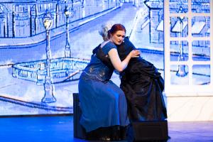 A woman in a blue dress sits on stage, embracing a person in black. The backdrop shows a blue-toned street scene, creating an emotional, theatrical atmosphere.