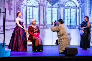 A stage scene with four actors in period costumes. One actor kneels to take a photo of two others. A backdrop shows large windows and ornate decor.