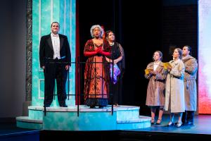 Five performers stand on a brightly lit stage, dressed in formal and vibrant attire, exuding a dramatic and emotive atmosphere against a colorful backdrop.