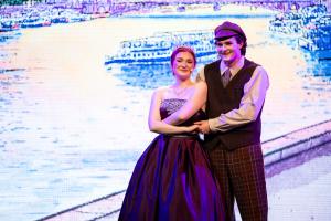 A woman in a gown and a man in vintage attire smile and pose in front of a painted river backdrop. The scene conveys a joyful, theatrical mood.