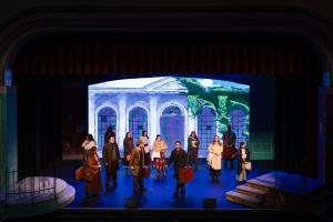 A theatrical performance on stage with actors in period costumes. The backdrop displays a grand building with arches, conveying a dramatic, historical setting.
