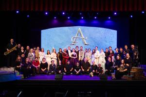 The cast of Anastasia poses on a stage under blue lighting. They are diverse in attire. The backdrop features a prominent letter "A."