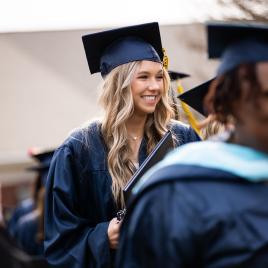 Women smiling in graduation regalia outside with another woman blurred in the foreground