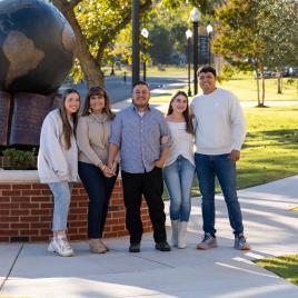 Family of five standing outside on a sidewalk in front of green trees. 