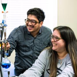 A male and female student in a science lab with goggles on smiling and using science beakers.