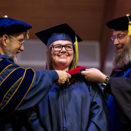 Two men on the sides and a woman in the center in academic regalia. The woman is being hooded