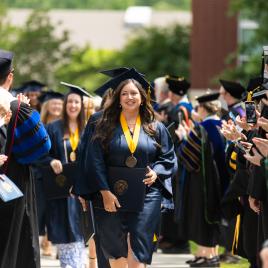 Graduates in navy caps and gowns walking through a tunnel of professors in academic regalia.