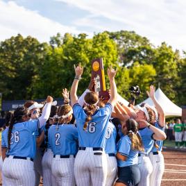 The ETBU women’s softball team in light blue jerseys and white pants holds up the NCAA Division III National Championship trophy. The scene is outdoors with green trees in the background. Several players raise their hands in celebration, while one person films the moment. 