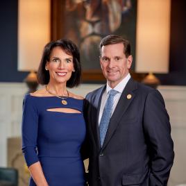 A woman in a blue dress and a man in a dark suit stand side by side, smiling in a warmly lit, formal room.
