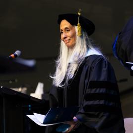 A woman in academic regalia standing on a stage with a dark background