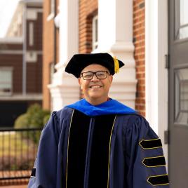 A man in doctoral regalia smiling in front of a red brick building with white columns.