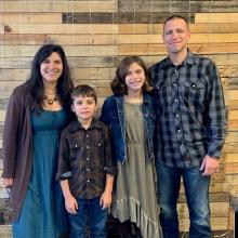 A family of four standing in front of a wooden plank wall, smiling.