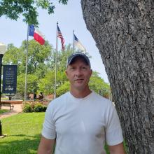 Man in a white shirt, standing next to a tree in a park with flags in the background.
