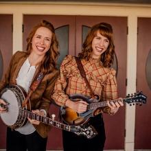 Two woman smiling playing banjos in front of red and black doors