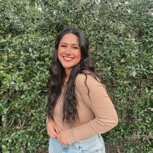 Woman in a tan longsleeve and jeans smiling at the camera with trees in the background