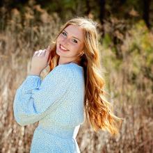 Caylee Mayfield Woman in a blue dress smiling at camera with tall grass in the background