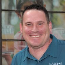 A person smiling in front of a window with blurred background elements.
