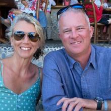 Smiling middle-aged couple wearing sunglasses, the woman in a blue polka-dot top and the man in a blue button-down shirt, seated together outdoors.