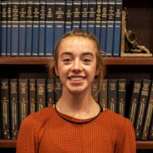 Girl in a orange shirt in front of a bookcase.