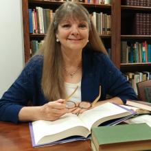 A woman with long brown hair is sitting at a table in a library, surrounded by open books. She is wearing a blue sweater and holding a pair of glasses. Bookshelves filled with books are in the background.