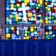 Four glass awards sit on a blue-draped table in front of a colorful stained glass window with a prominent white cross.