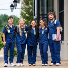Five nursing students in blue scrubs stand together outside a building, smiling with their arms around each other. They are wearing backpacks and have identification badges, with stethoscopes and books visible.