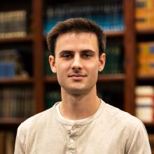 A young man with short dark hair wearing a light beige Henley shirt stands in front of a bookshelf filled with books, looking at the camera with a neutral expression.
