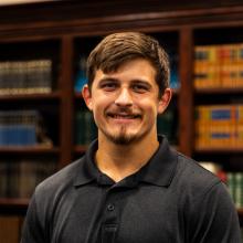 A young man with short brown hair and a mustache smiles while standing in front of a bookshelf filled with books.