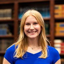 A young woman with long blonde hair wearing a bright blue shirt smiles at the camera, standing in front of a bookshelf filled with books.