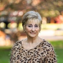 A woman with short, light blonde hair, wearing a fuzzy leopard-print sweater, stands outdoors in a natural, blurred background setting.