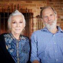 Elderly couple sitting together indoors, with a brick wall and iron detailing in the background.