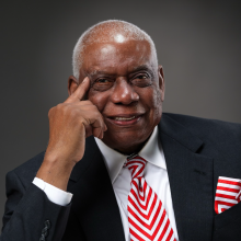 moore Elderly man in a dark suit with a red and white chevron patterned tie and matching pocket square, smiling subtly.