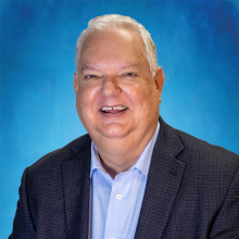 terrell Portrait of a smiling older man with short white hair, wearing a light blue shirt and dark gray blazer, against a bright blue background.